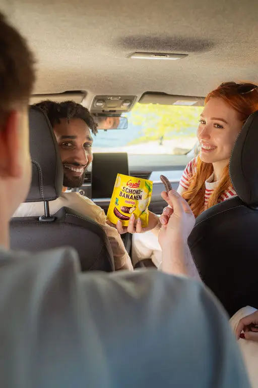 Junge Leute sitzen im Auto und teilen sich eine Packung Schoko-Bananen Minis