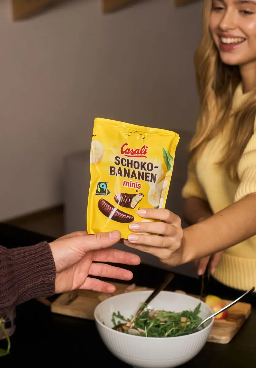 Jemand gibt einer jungen blonden Frau eine Packung Schoko-Bananen Minis